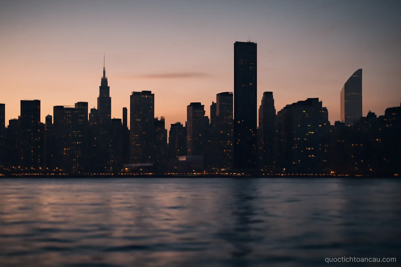 a dense city skyline at dusk seen across water with lights beginning to come on